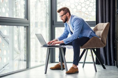 İyi bakımlı sakallı ve gözlüklü yakışıklı erkek ofis müdürü, modern ofis merkezinde ayrı yerlerde dizüstü bilgisayar ve mali raporlar kullanıyor..