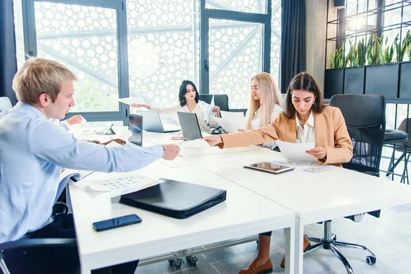 Compañeros de carreras mixtas analizando diferentes papeles de negocios y trabajando con computadoras en las mesas de la oficina moderna . — Foto de Stock