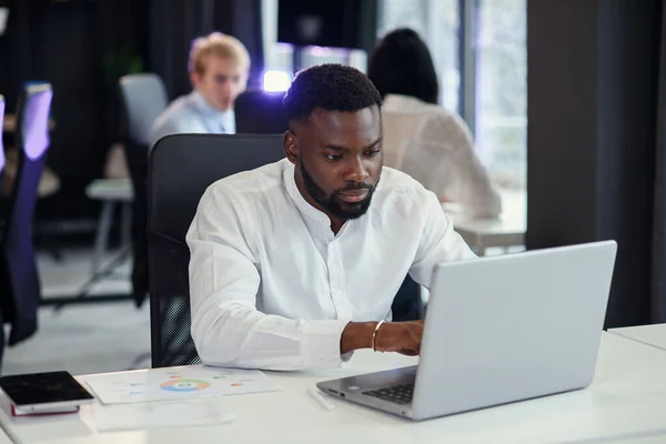 Serieuze jonge kantoormanager met een donkere huid werkt met verschillende rapporten en tafellaptop op zijn werkplek in de vergaderruimte van het moderne businesscentrum. — Stockfoto