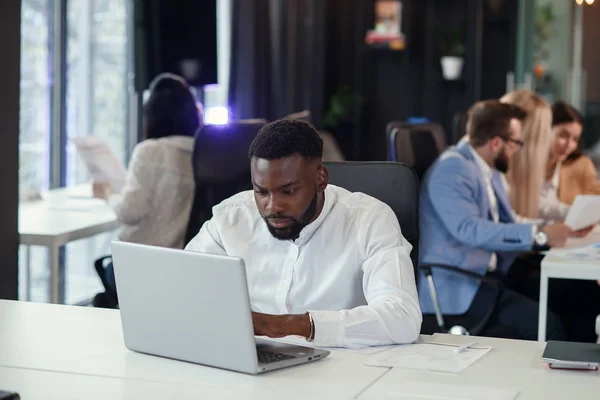 Serieuze jonge kantoormanager met een donkere huid werkt met verschillende rapporten en tafellaptop op zijn werkplek in de vergaderruimte van het moderne businesscentrum. — Stockfoto