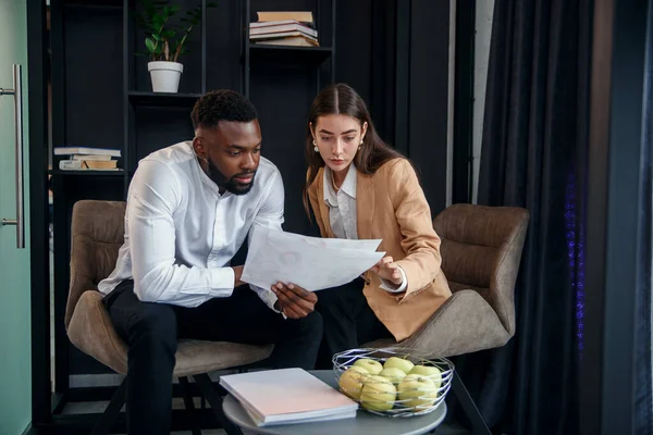 Pareja de negocios de hombre afroamericano y mujer europea que se sientan en una habitación especialmente designada y utilizando documentos en papel y tableta PC, conversando sobre su proyecto empresarial y su — Foto de Stock