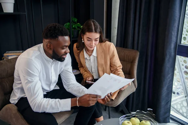 Pareja de negocios de hombre afroamericano y mujer europea que se sientan en una habitación especialmente designada y utilizando documentos en papel y tableta PC, conversando sobre su proyecto empresarial y su — Foto de Stock