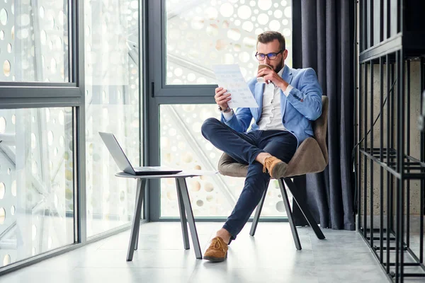 Fiatal sikeres üzletember elemzi a kezében lévő pénzügyi dokumentumot, és illatos kávét iszik a modern coworking center speciálisan kijelölt szobájában.. — Stock Fotó