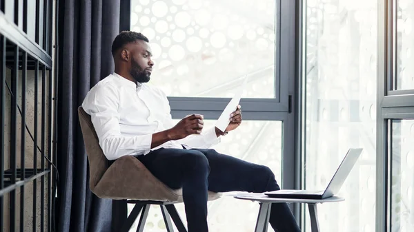 Hombre de negocios afroamericano guapo que trabaja en la computadora portátil y el análisis en la habitación especialmente designada en la oficina moderna y elegante . — Foto de Stock