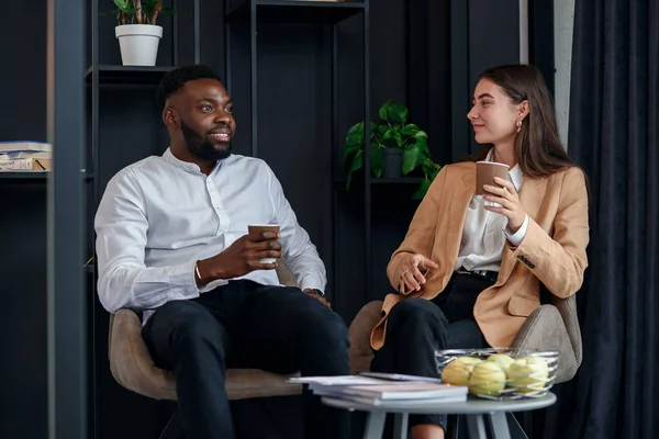 African man and caucasian woman have coffee break at office rest room at table near window drinking hot espresso.