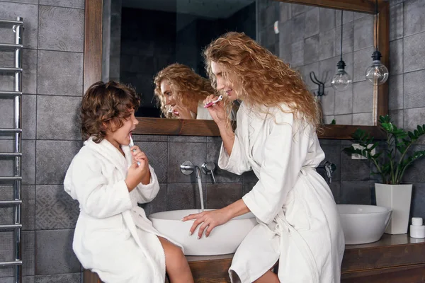 Krásná matka s blond kudrnaté vlasy v bílém županu čištění zubů s její roztomilý kudrnatý syn v ranní přední části zrcadla v moderní koupelně. — Stock fotografie