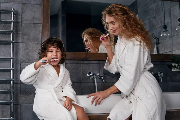 Γοητευτική μαμά με λευκό φόρεμα με σγουρά μακριά μαλλιά διδάσκει χαριτωμένο μικρό γιο της να βουρτσίζει τα δόντια στο μπάνιο το πρωί. — Φωτογραφία Αρχείου
