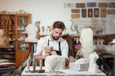 Sakallı zanaatkar bir keskiyle beyaz taş oymacılığında çalışıyor. Sanat eserleri ile yaratıcı atölye.