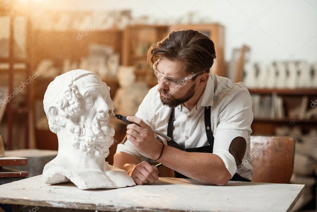 Male sculptor repairing gypsum sculpture of womans head at the working place in the creative artistic studio.