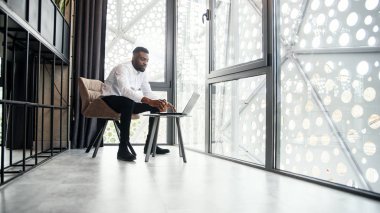 Laptopta çalışan yakışıklı Afro-Amerikan iş adamı ve modern şık ofisin özel olarak belirlenmiş odasında analizler yapıyor..