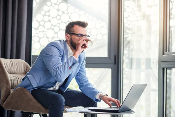 Ung framgångsrik affärsman analyserar finansiella dokument i handen och drycker doftande kaffe i särskilt utsedda rum i moderna coworking center. — Stockfoto