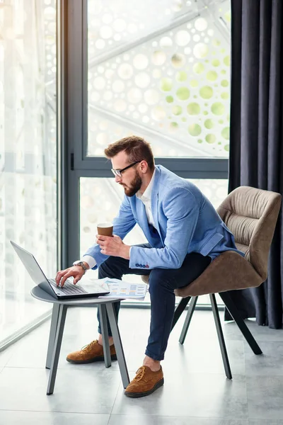 Fiatal sikeres üzletember elemzi a kezében lévő pénzügyi dokumentumot, és illatos kávét iszik a modern coworking center speciálisan kijelölt szobájában.. — Stock Fotó