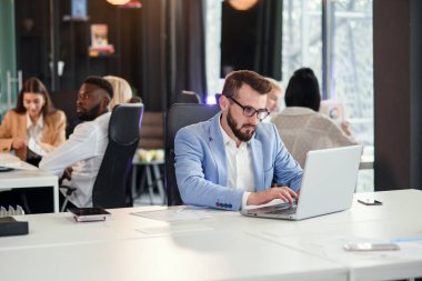 Gözlüklü ve mavi takım elbiseli başarılı sakallı işadamı modern ofiste dizüstü bilgisayarla çalışıyor..