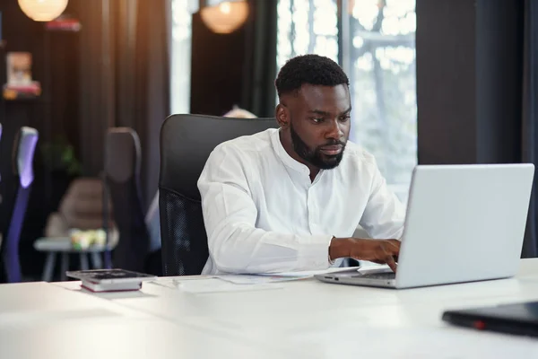 Serieuze jonge kantoormanager met een donkere huid werkt met verschillende rapporten en tafellaptop op zijn werkplek in de vergaderruimte van het moderne businesscentrum. — Stockfoto