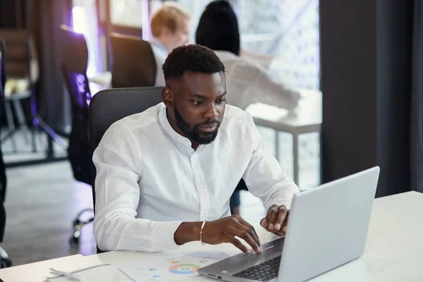 Serieuze jonge kantoormanager met een donkere huid werkt met verschillende rapporten en tafellaptop op zijn werkplek in de vergaderruimte van het moderne businesscentrum. — Stockfoto