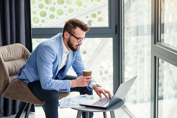 Close up of industrious good-looking young businessman analising document in his hand and drinking fragrant coffee in specially designated room in modern office