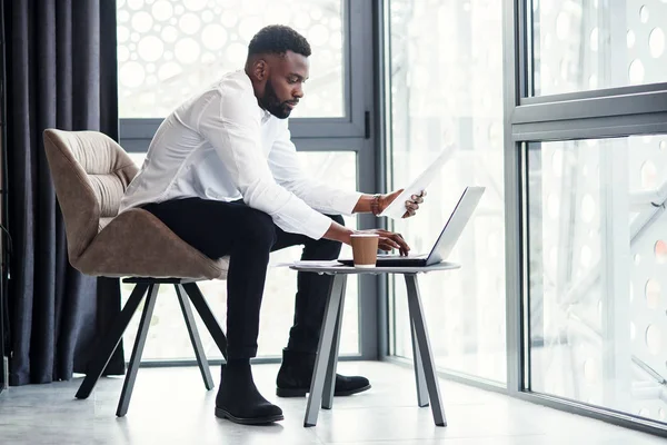 De jonge Afrikaanse zakenman zit aan het bureau met een laptop. Guy kijkt naar papieren documenten en officiële rapporten. — Stockfoto