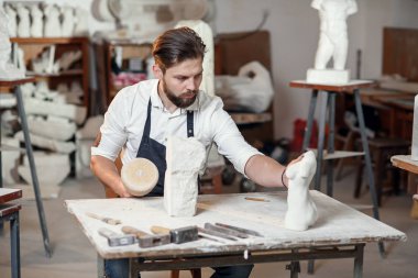 Beyaz gömlekli ve siyah önlüklü erkek heykeltıraş sanat stüdyosunda kadın gövdesinin kireçtaşından bir kopyasını yapıyor..