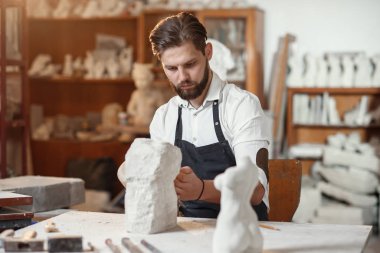 Beyaz gömlekli ve siyah önlüklü erkek heykeltıraş sanat stüdyosunda kadın gövdesinin kireçtaşından bir kopyasını yapıyor..