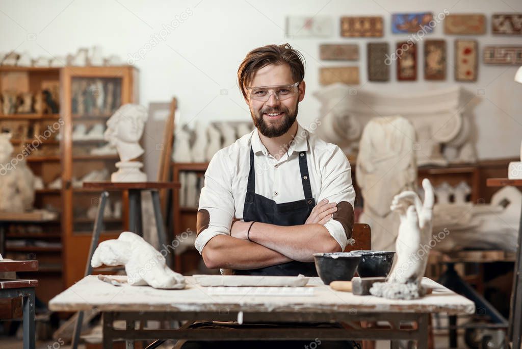Portrait of attractive joyful bearded male sculptor which sitting on the workshop interior background and looks at the camera with smile.