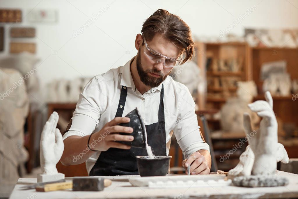 Unrecognizable professional stonemason with well-maintained hands making glue in special vessel for his handmade sculpture