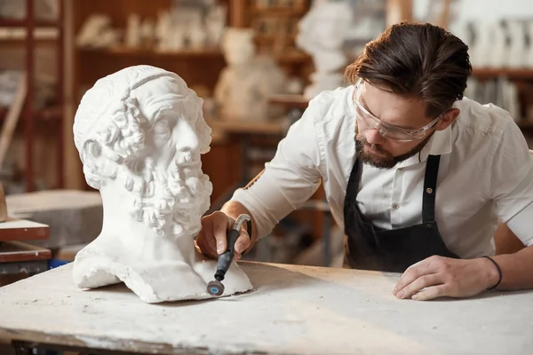 Male sculptor repairing gypsum sculpture of womans head at the working place in the creative artistic studio.