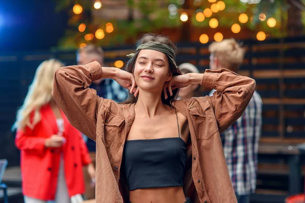 Menina bonita dançando em primeiro plano, enquanto seus amigos dançando à luz de lâmpadas coloridas durante a festa de celebração ao ar livre . — Fotografia de Stock