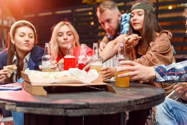 Amigos multi-étnicos desfrutando de pizza e cerveja na festa. Jovens a dar uma festa. Foco seletivo em garrafas de bebidas energéticas à mesa . — Fotografia de Stock