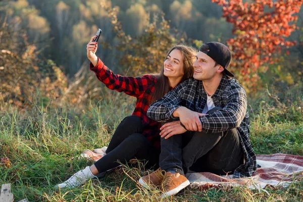 Genç mutlu hipster çift, bahar parkında çimlerde otururken akıllı telefon kullanarak selfie çekiyor. Modern çiftler, doğada oturan arkadaşlarıyla bir video görüşmesi yaparlar.. — Stok fotoğraf