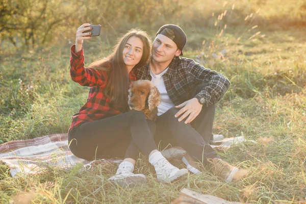 Snygga par har ett videosamtal med sina vänner. Young Happy hipster par med sin hund att göra Selfie med hjälp av smart telefon när du sitter på gräsmattan i Spring Park. — Stockfoto