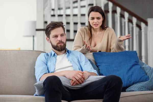 Confused man sits on the couch while his girlfriend yells and quarrels with him at home. Big family problems. — Stock Photo, Image