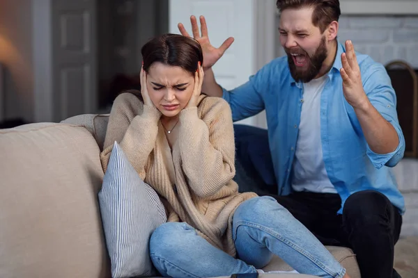 Defenseless upset woman closes her ears while her husband yelling at her sitting behind. Family relationship problems concept. — Stock Photo, Image