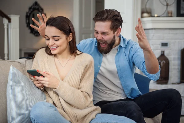 Young wife uses smartphone while sitting on sofa and her husband feels upset behind she. Problems in the family due to addiction to the smartphone.