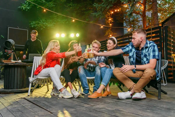 Amigos clinking garrafas e beber cerveja ao ar livre festa aconchegante com música e luzes de discoteca coloridas . — Fotografia de Stock