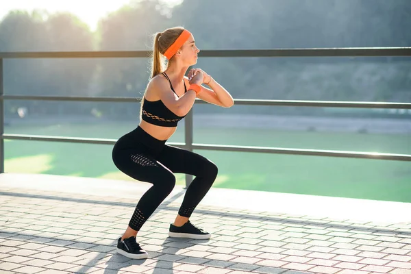 Preciosa chica deportiva en ropa deportiva negra y diadema naranja que se agacha durante el entrenamiento de fitness en el estadio al aire libre . —  Fotos de Stock