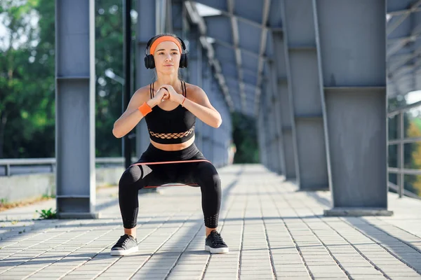 Chica de fitness con auriculares que hacen ejercicios en cuclillas con banda de tejido botín durante su entrenamiento deportivo en un campo de deportes especial — Foto de Stock