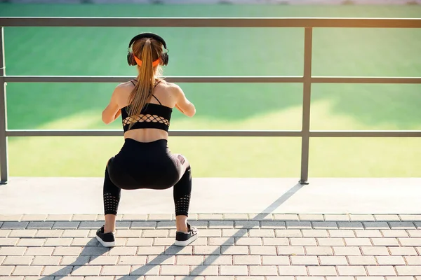Fitness girl with earphones which doing squat exercises with fabric booty band during her sport workout on special sportground — Stock Photo, Image