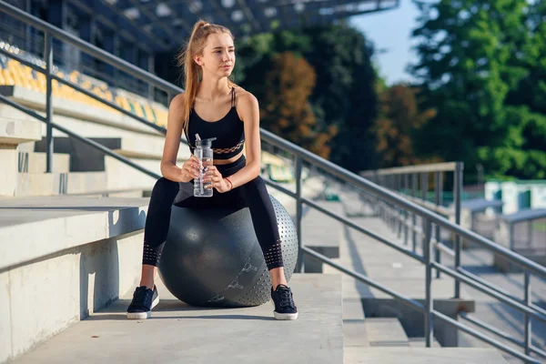 Νεαρή αθλητική γυναίκα σε κομψό μαύρο αθλητικό ντύσιμο κρατώντας ένα μπουκάλι νερό στο χέρι, ενώ κάθεται στην μπάλα γυμναστικής. Εξαντλημένη, όμορφη λεπτή κορίτσι γυμναστικής έχουν ανάπαυση και πόσιμο νερό μετά την προπόνηση. — Φωτογραφία Αρχείου