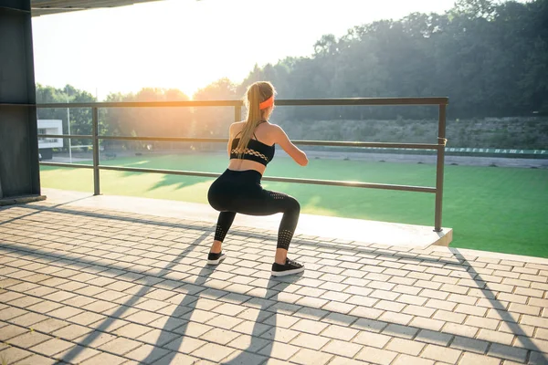 Preciosa chica deportiva en ropa deportiva negra y diadema naranja que se agacha durante el entrenamiento de fitness en el estadio al aire libre . — Foto de Stock