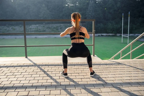 Preciosa chica deportiva en ropa deportiva negra y diadema naranja que se agacha durante el entrenamiento de fitness en el estadio al aire libre . — Foto de Stock