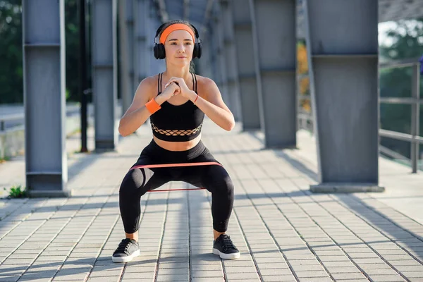 Fitness girl with earphones which doing squat exercises with fabric booty band during her sport workout on special sportground