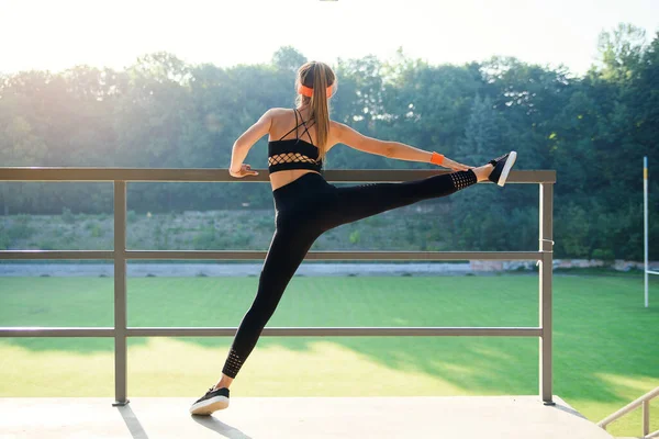 Muchacha atractiva en ropa deportiva haciendo ejercicios de estiramiento para piernas en el estadio de la ciudad. Mujer de fitness haciendo ejercicio de calentamiento al aire libre . — Foto de Stock