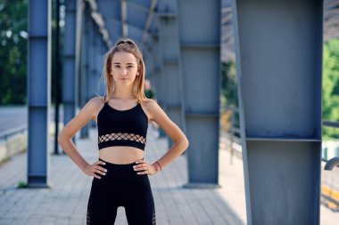 Spor kıyafetli, genç, beyaz, sarışın bir kız kalçalarının üstünde el ele tutuşuyor, poz veriyor ve kameraya bakıyor..