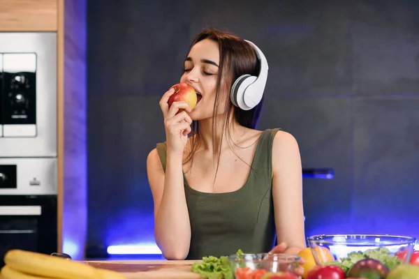 Young attractive woman eats red apple and listens to the music at kitchen in morning. Healthy lifestyle concept, enjoying breakfast.