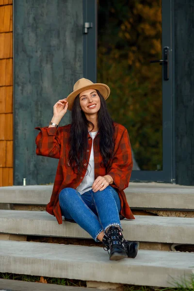 Jovem mulher em um chapéu marrom camisa vermelha na moda e calças jeans senta-se em uma escada, perto de sua própria casa . — Fotografia de Stock