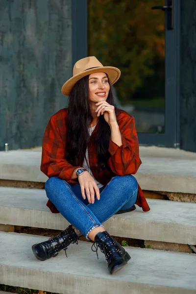 Schöne Frau in rotem Hemd, Jeans und Hut sitzt auf der Treppe ihres modernen Hauses in der Nähe des Waldes. — Stockfoto