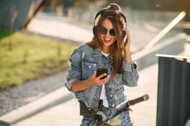Yazlık kıyafetler giyen güzel, gülümseyen genç bir kız şehir merkezindeki bir bisikletin üzerinde oturur ve cep telefonlu kablosuz kulaklıklarla müzik dinler. Modern akıllı teknoloji.