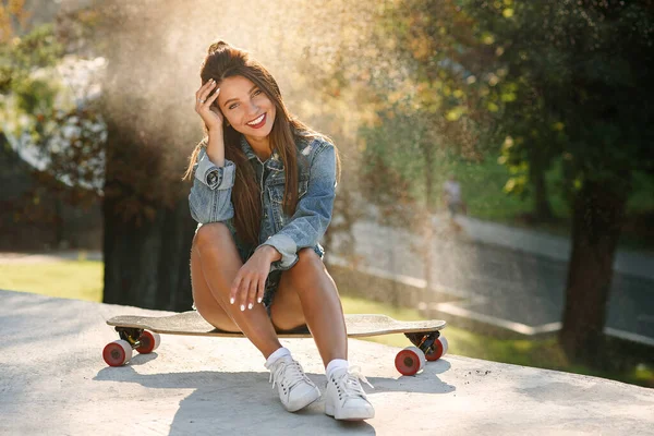 Bela jovem mulher alegre senta-se no skate no espaço urbano ao nascer do sol . — Fotografia de Stock