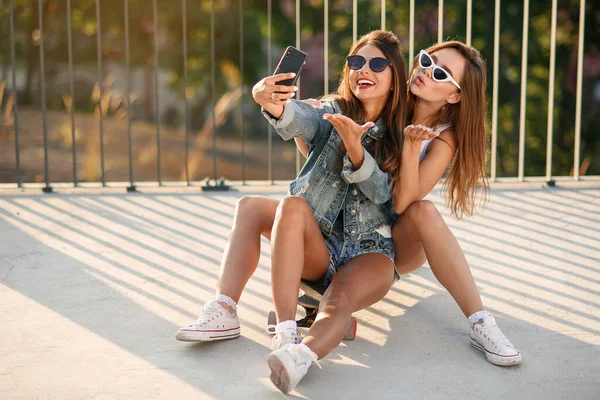 Dos novias adolescentes en traje hipster sentadas en el monopatín en el parque y hacer foto selfie en el teléfono inteligente. Feliz amistad y estilo de vida activo . —  Fotos de Stock