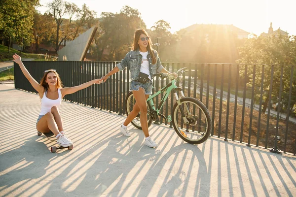 Caucasiano menina bonita senta-se em um longboard enquanto sua namorada vai para a frente e segura a mão . — Fotografia de Stock